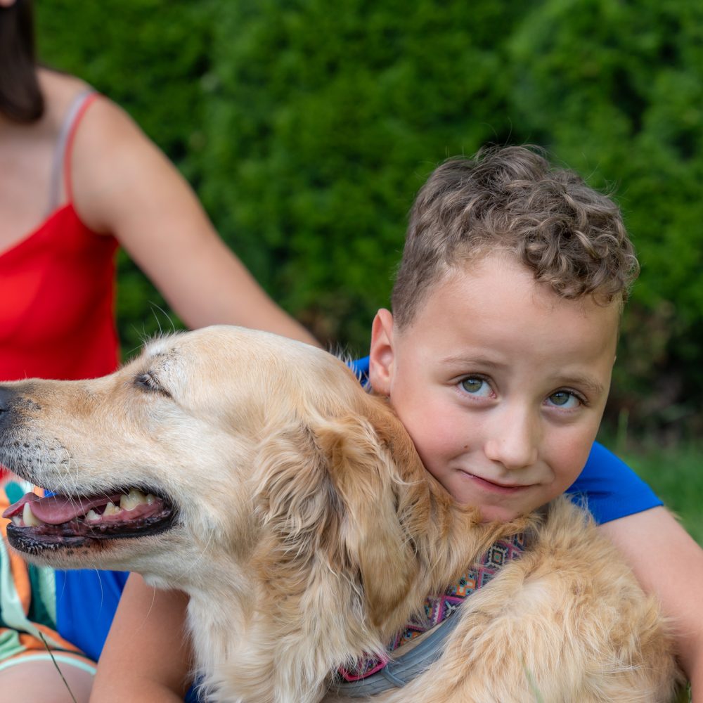 Young Boy Gently Hugs A Calm Golden Retriever While His Mother Sits Nearby In A Green Garden. The Warm Outdoor Scene Reflects Family Love, Pet Companionship, Trust, Childhood Happiness, And Animal Care.